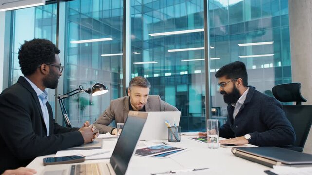 Business team in formal wear having focused discussion at meeting table in modern office. Man in grey blazer leading conversation, others listening attentively with laptops and data sheets.
