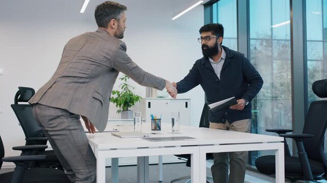 Caucasian man in suit shaking hands with South Asian man during job interview. Confident gesture at meeting in modern office. CV or resume reading. Interviewing for new work position in company.