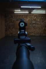 First-person shooter perspective aiming a modern rifle with a collimator red dot sight down an indoor tactical shooting range.