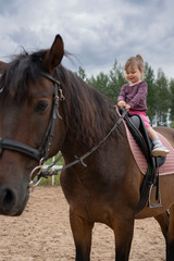 Fototapeta premium Cute little girl riding a horse outdoors. Pet therapy. Hippotherapy for young children, therapy after many serious diseases.