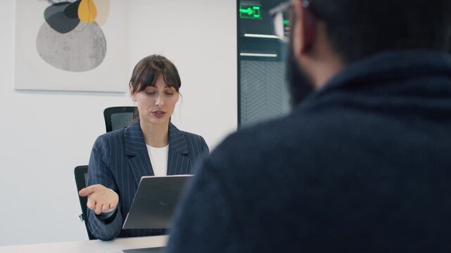Caucasian HR woman holding folder with CV, interviewing man during hiring process. Office decorated with modern art and white walls. Serious expression, focused discussion. Interview for job concept.