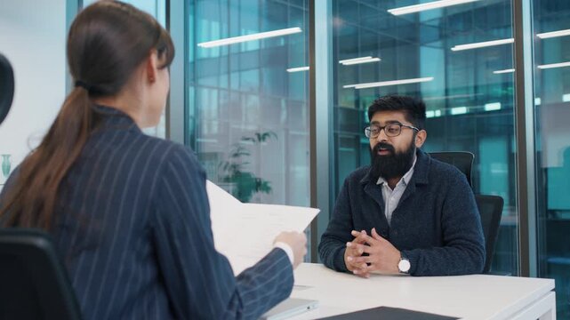 Indian man with beard and glasses sitting confidently during interview, Caucasian HR woman holding resume, discussing qualifications in modern glass office. Female boss interviewing new employee.