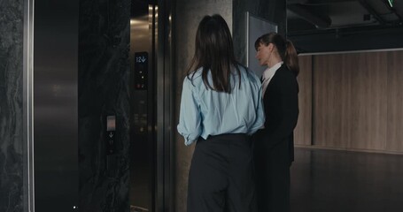 Two professional office workers girls call the elevator and enter it while chatting. Office girls in business clothes chatting and entering the elevator