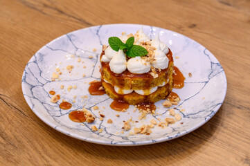 Gourmet dessert with caramel, nuts, sponge cake, and whipped cream on a marble plate, served on a rustic wooden table in fine dining restaurant setting.