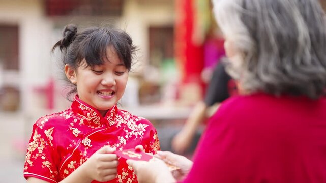 Chinese lunar new year traditional holiday festive event celebration. Happy Asian family grandmother giving money gift in red envelopes and blessing to little grandchild girl in Chinese red dress.