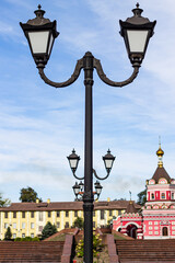 A classic twin lamppost stands tall in the foreground against a historical cityscape with an ornate church and buildings under a bright blue, cloudy sky, suggesting a public square.


