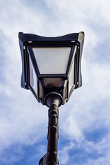 A low-angle shot of a classic black street lamp (lantern) against a bright blue sky with wispy clouds. The design suggests vintage or historical urban architecture.

