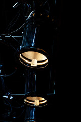 Two theatrical spotlights with warm, golden illumination hang vertically in absolute darkness. The scene is dramatic and moody, highlighting professional stage equipment.

