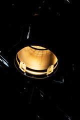 A dramatic close-up of a warm, golden stage light (spotlight) emerging from deep darkness. The light source creates a moody, intense atmosphere with sharp contrast.

