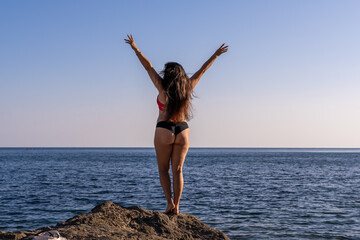Woman ocean freedom, young lady celebrating summer vacation on rocky beach with arms raised, blue sky copy space