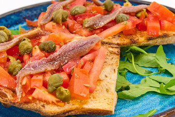 Italian bruschetta with fresh tomatoes, capers, anchovies and crusty bread on a blue plate