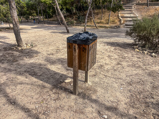 Wooden trash bin with black plastic bag, situated in a natural park setting surrounded by trees and dry earth, emphasizing environmental cleanliness and waste management