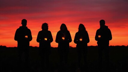 Five silhouetted figures hold candlelit objects against a vibrant red and orange sunset