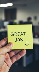 Great Job Note in Hand: Capturing the essence of encouragement and appreciation, a hand holds a yellow sticky note with the words GREAT JOB and a smiling face in an office environment.