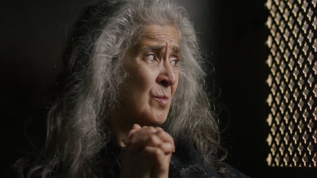 Faithful senior woman with long grey hair clasping her hands in prayer while sitting inside wooden confessional booth in church