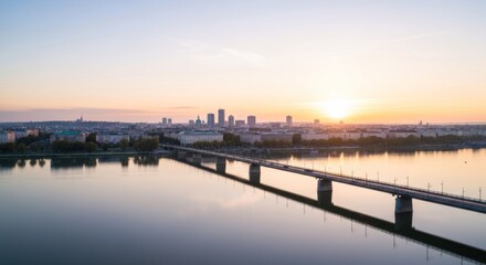 Obraz premium Panoramic city sunrise over river. Calm morning light reflects on water, a bridge stretches across. Modern buildings line the horizon