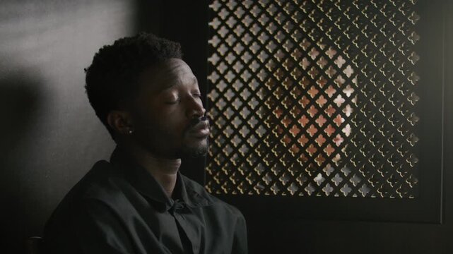 Young African American man sitting in confessional booth with ornate partition screen, answering questions from priest during confession