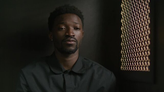 Young African American penitent sitting in dimly lit confessional booth near lattice screen, talking remorsefully to priest during confession