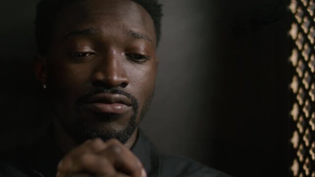 Young remorseful Black man sitting in dimly lit booth with lattice screen, holding hands clasped together as confessing his sins. Close-up view