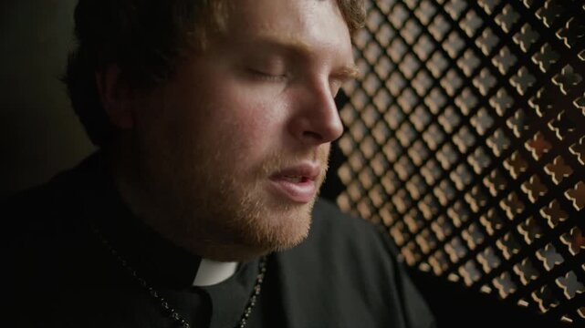 Close-up view of priest wearing black cassock and clerical collar talking to penitent through lattice partition inside confessional booth in church