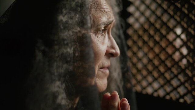 Senior woman in black veil holding hands clasped in prayer as speaking to priest through lattice screen during confession
