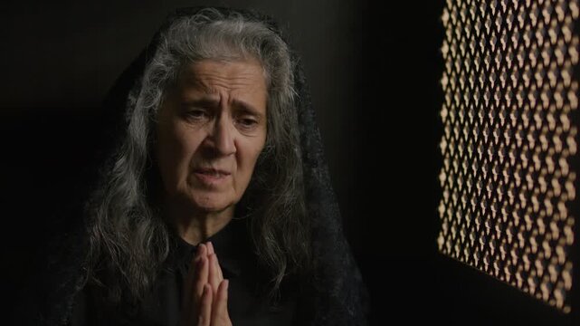 Elderly woman in black mantilla holding her hands together in prayer and talking sincerely to priest through confessional screen