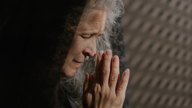 Senior woman in black lace veil praying deeply to God with her eyes closed and head bowed to clasped hands in confessional booth. Close-up profile view