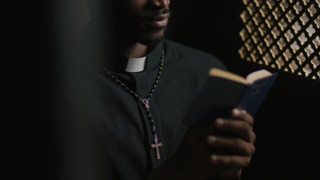 Black priest wearing rosary and clerical collar reading scripture aloud from small Bible near ornate screen in confessional. Close-up view