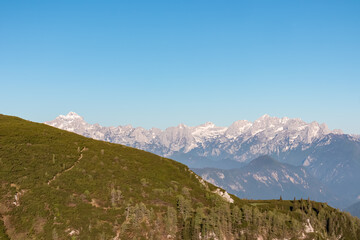 A narrow hiking trail crosses a green alpine meadow on Weinasch, offering a stunning panoramic view of the majestic Triglav and the snow-capped Julian Alps in Slovenia's Karawanks mountain range.