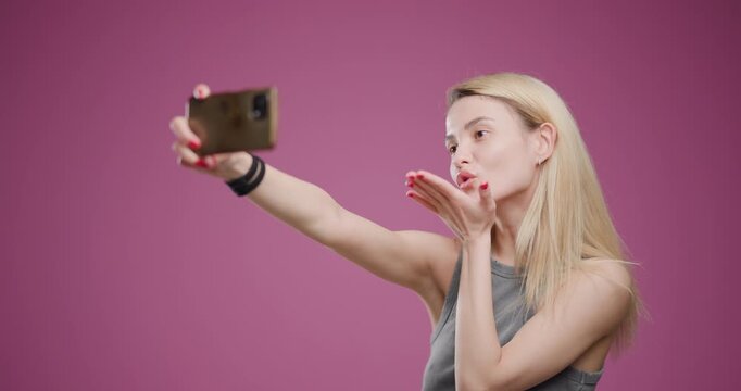 Young blonde woman taking a playful selfie, puckering lips on a pink background