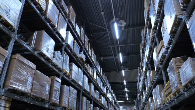 Interior of a large. Modern logistics center showing a long aisle with high metal racks filled with neatly stacked cardboard boxes and parcels ready for shipping. Representing supply chain management