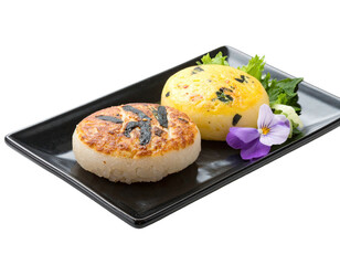 Two round, Japanese-style rice cakes, one browned, one yellow, on a dark plate, with purple flowers