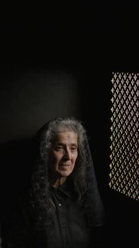 Elderly female parishioner in black veil sitting inside confessional booth, speaking through patterned screen beside her. Vertical clip