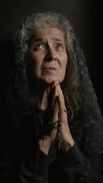 Elderly female parishioner in black veil holding rosary beads, clasping hands in deep prayer inside dark confessional booth. Vertical format video