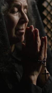 Senior woman seen holding hands together in deep prayer with rosary wrapped around fingers inside dark confessional booth. Vertical clip, close-up view
