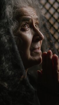 Senior woman in black veil whispering prayer with eyes closed and rosary in her clasped hands during spiritual confession. Vertical close-up shot