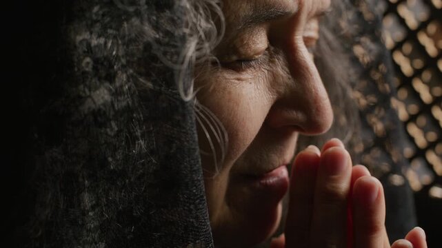 Elderly woman in dark mantilla holding hands near lips and whispering prayer during confession. Close-up view