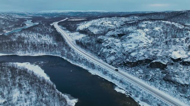 Murmansk, Russia - December 13, 2024: Aerial view of a black Jetour T2 SUV driving on a snowy road situated between a partially frozen river and a steep, snow-dusted, rocky hill in the arctic valley
