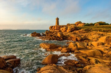 Golden hour view of a lighthouse on the Pink Granite Coast in Brittany, France, with dramatic orange rocks and waves crashing against the rugged shoreline under a serene sky.