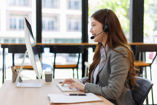 Happy young asian woman Call Center Person wearing a headset, providing support at a call center, smiling while typing on computer keyboard
