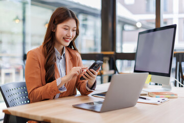 Asian businesswoman working using phone and laptop computer at her desk in a bright modern office, doing calculating expense financial report finance making notes paper graph data document.	