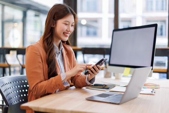 Asian businesswoman working using phone and laptop computer at her desk in a bright modern office, doing calculating expense financial report finance making notes paper graph data document.	 - Powered by Adobe