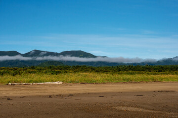 A coastal plain with grasses and sand lies below a dark green mountain ridge, where a layer of mist drifts across under clear blue sky. Nature of Sakhalin Island