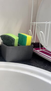 A sink with a white plate on it and a sponge holder with two sponges in it.