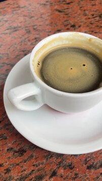 A cup of coffee sits on a white plate on a red table. The coffee is dark and has a strong aroma. The cup is almost empty, with only a small amount of coffee left in it