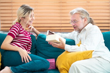 Smiling grandfather and granddaughter with gift boxes sitting on couch at home. Senior father with young woman indoors at home at Christmas, Holding present.