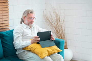 Senior man using digital tablet on sofa. Retired man enjoy playing computer game. Searching data. Online shopping. Watching movie or video call with family at home