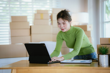 A young Asian female entrepreneur stands in her home office holding a tablet and a parcel box to prepare to manage online orders and deliveries.