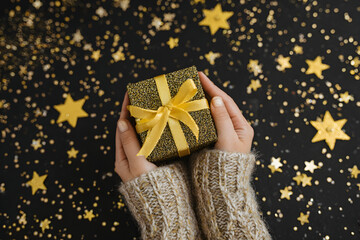 Hands holding a golden glittery gift box with a yellow ribbon on a star speckled black background