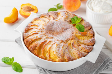 Delicious peach pie in a white ceramic baking form on a white wooden background. Summer cake. Selective focus, copy space.